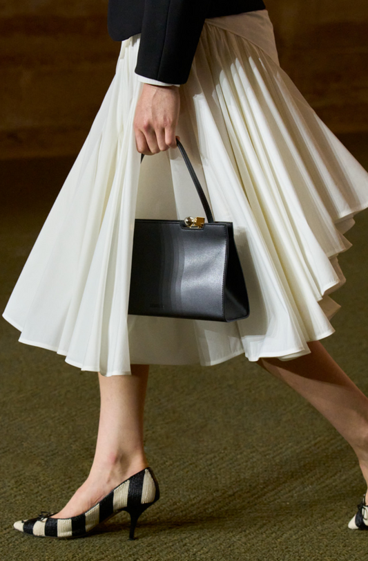 Person wearing a white pleated skirt and black high-heeled shoes, holding a black handbag.